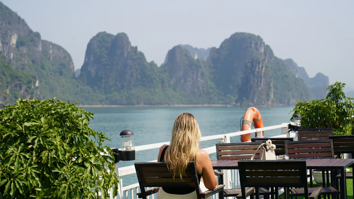 Woman with long hair sits at a wooden table on a boat, gazing at limestone islands over calm water. Bright, sunny day with lush greenery.