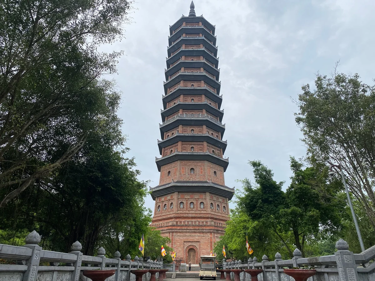 Tall pagoda with intricate brickwork stands between lush green trees under a cloudy sky, flanked by stone railings and small flags.