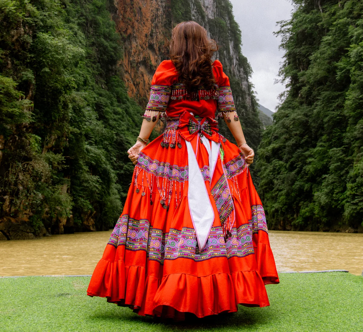 Woman in vibrant red traditional dress stands before a river, surrounded by lush green cliffs, appreciating the natural scenery.