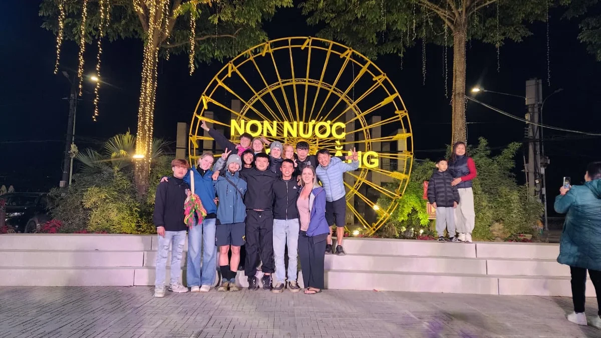 Group of people posing happily at night in front of a lit yellow structure with "NON NUOC" text. Trees and lights decorate the background.