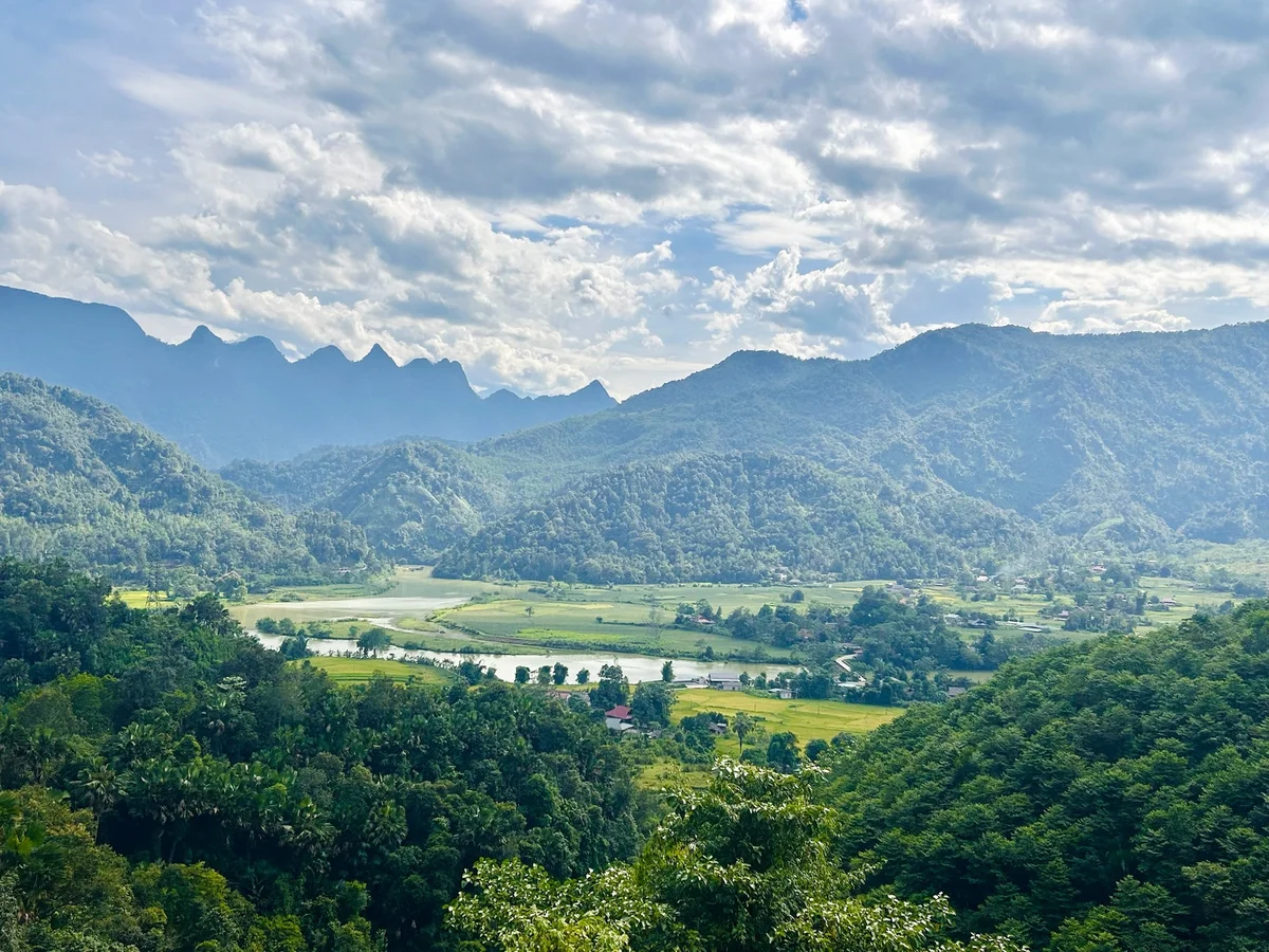 Lush green valley with a winding river, surrounded by forested mountains under a partly cloudy sky, creating a serene landscape.