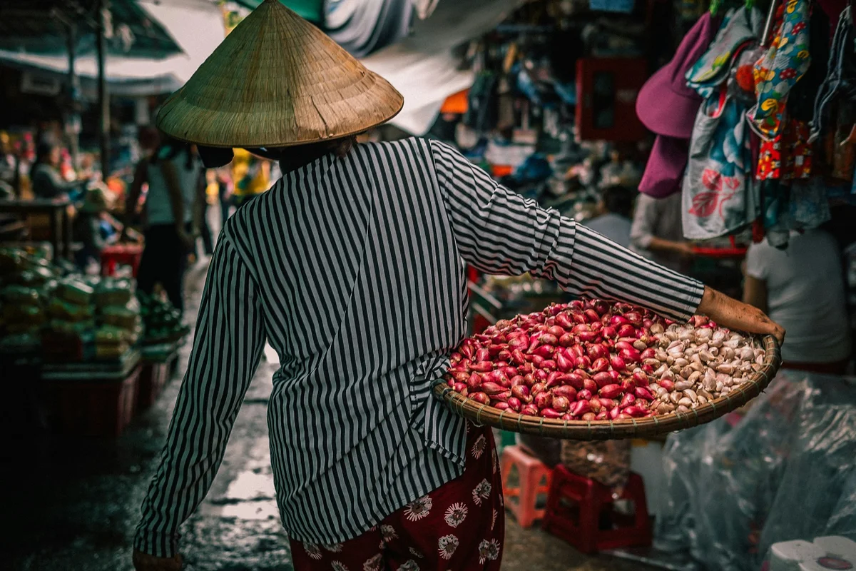 A person in a striped shirt and conical hat carries a basket of red onions in a bustling, colorful market setting.