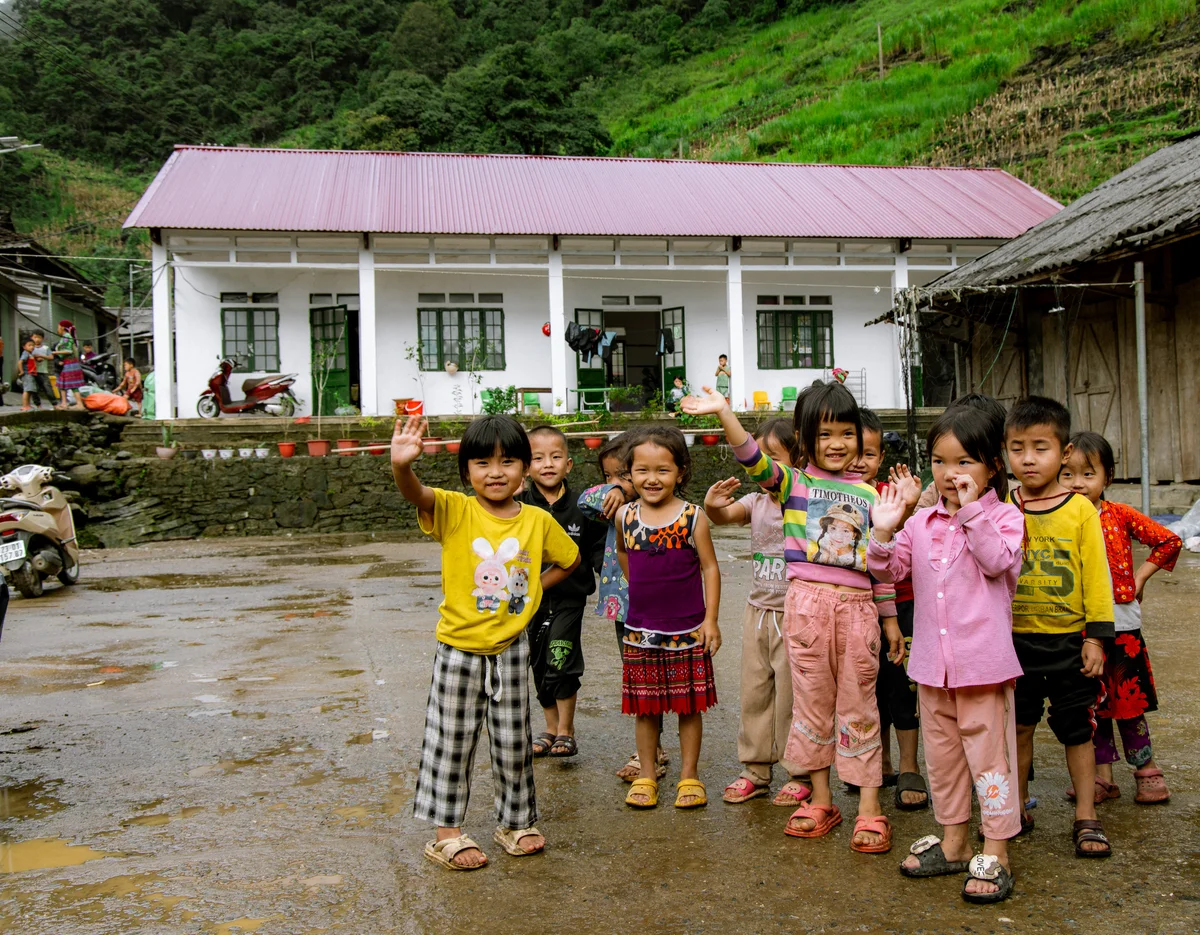Group of children smiling and waving outside a white house with a red roof. Green hills in the background. Cheerful, vibrant scene.