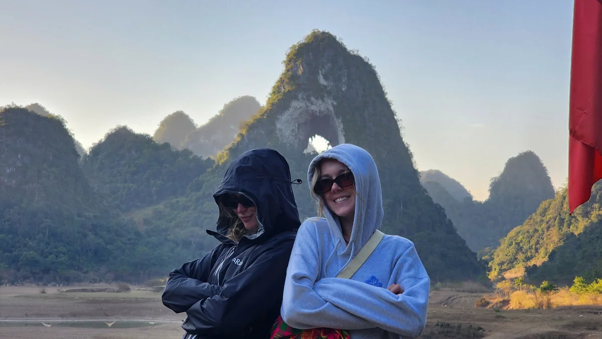 Two people in hoodies and sunglasses pose with arms crossed in front of scenic mountains, under a clear sky. A red flag is on the right.