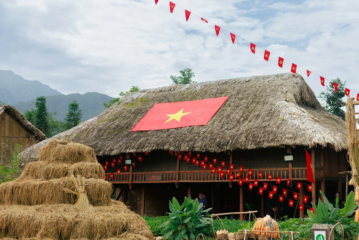 Thatch-roofed house displaying a large red Vietnam flag. Red pennants feature yellow stars. Mountainous backdrop with haystack and greenery.