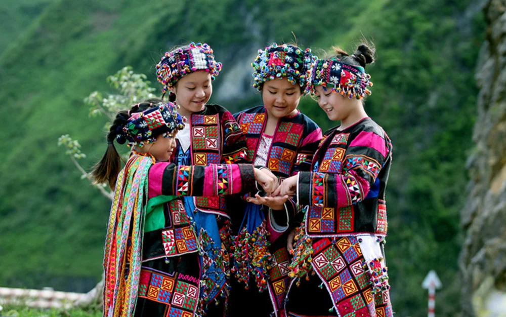 Four children in colorful traditional attire, adorned with beaded headdresses, stand in a green landscape, smiling and interacting.