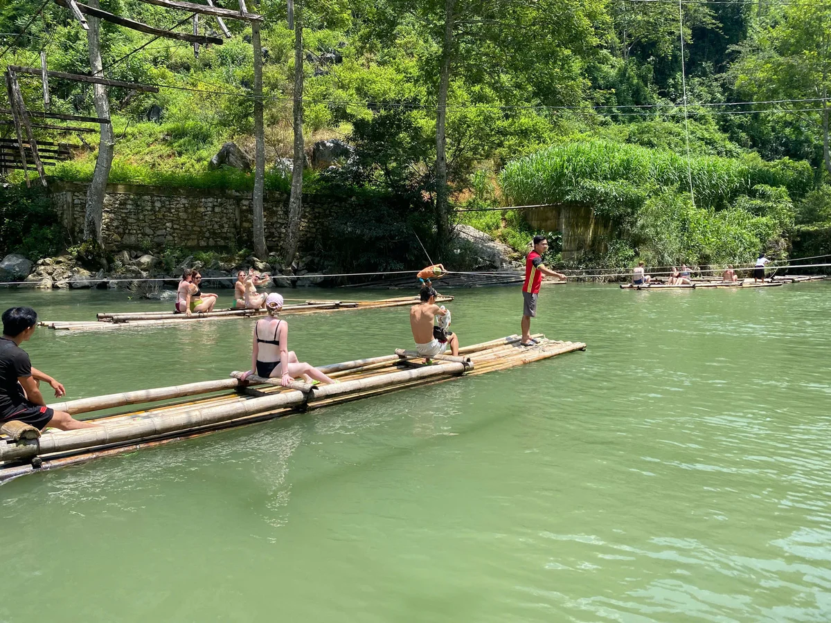 People on bamboo rafts relax and chat on a green river, surrounded by lush trees and a stone wall. Sunny day, calm and peaceful setting.