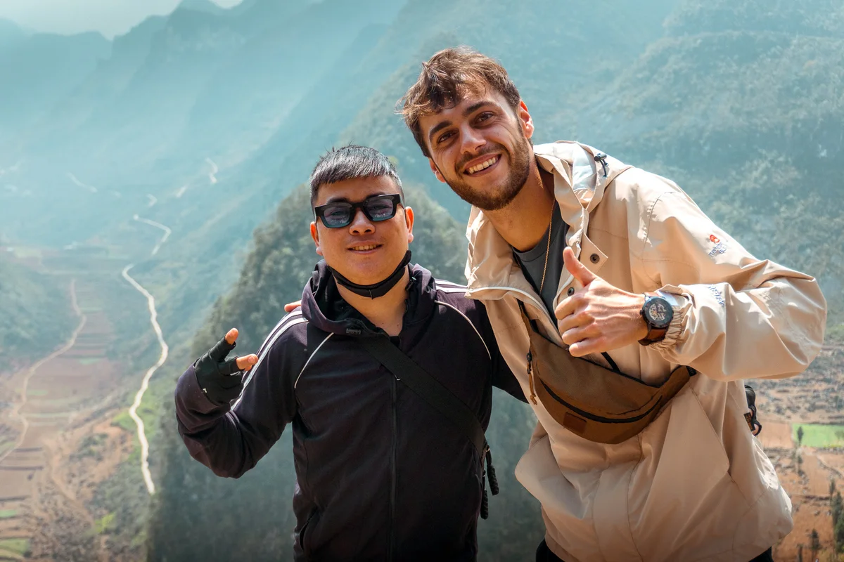 Two smiling men pose with thumbs up and peace sign in front of mountains under a cloudy sky. One wears sunglasses, the other a watch.