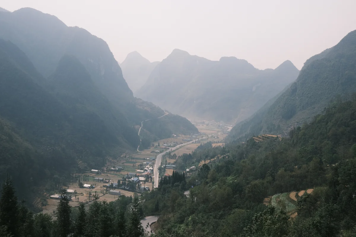 Mountain valley view with lush green hills surrounding a winding road and scattered village houses. Misty atmosphere under a pale sky.
