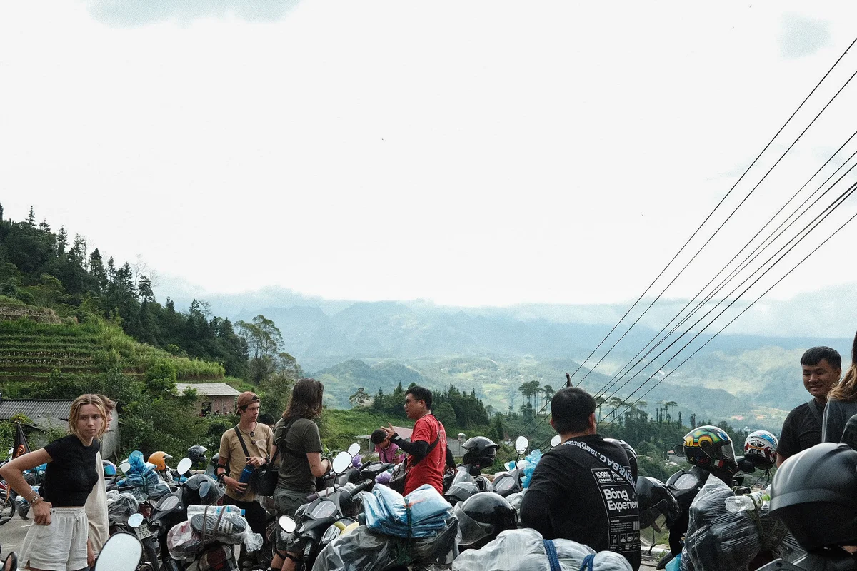 People standing around lots of motorbikes and helmets and mountains behind