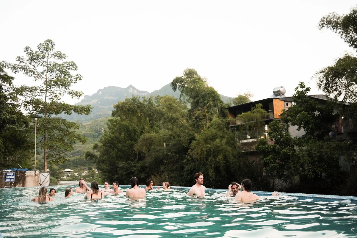 People enjoying a pool surrounded by trees and mountains, with modern buildings nearby. The mood is relaxed and social.