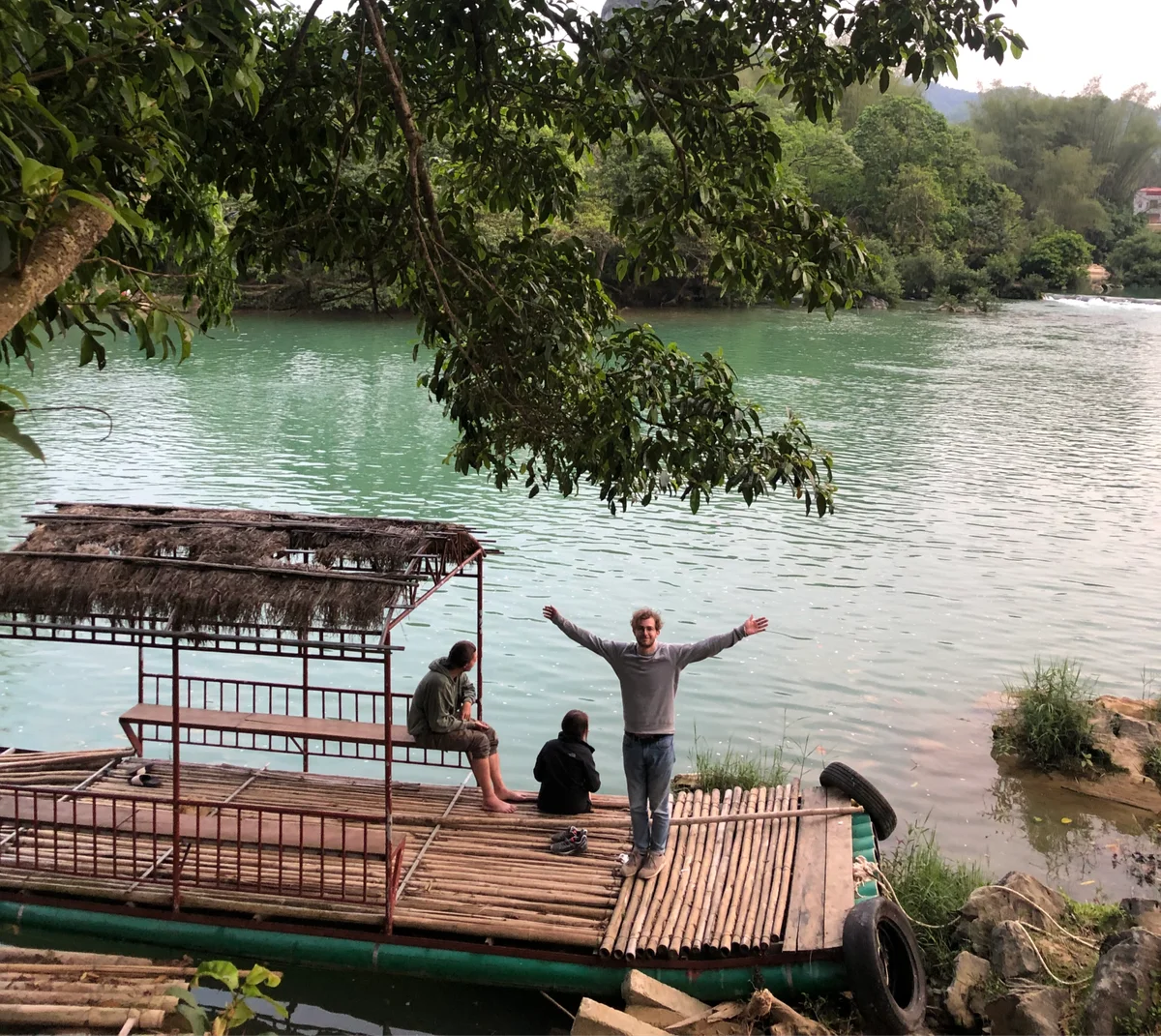 Three people on a bamboo raft by a serene, green river. One person stands with arms outstretched, two sit. Lush trees surround the scene.