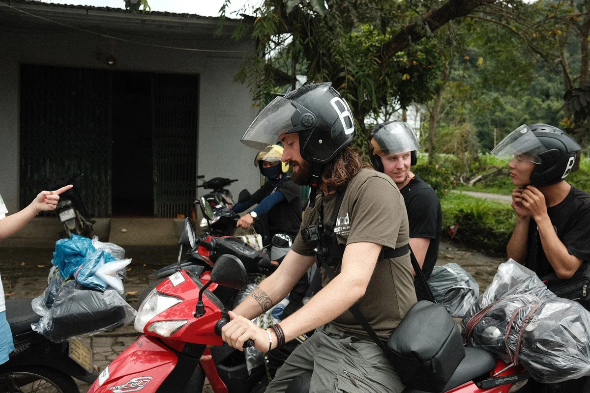 Group of people on motorbikes wearing helmets, gathered outdoors near a building. A person is pointing, and there's greenery in the background.