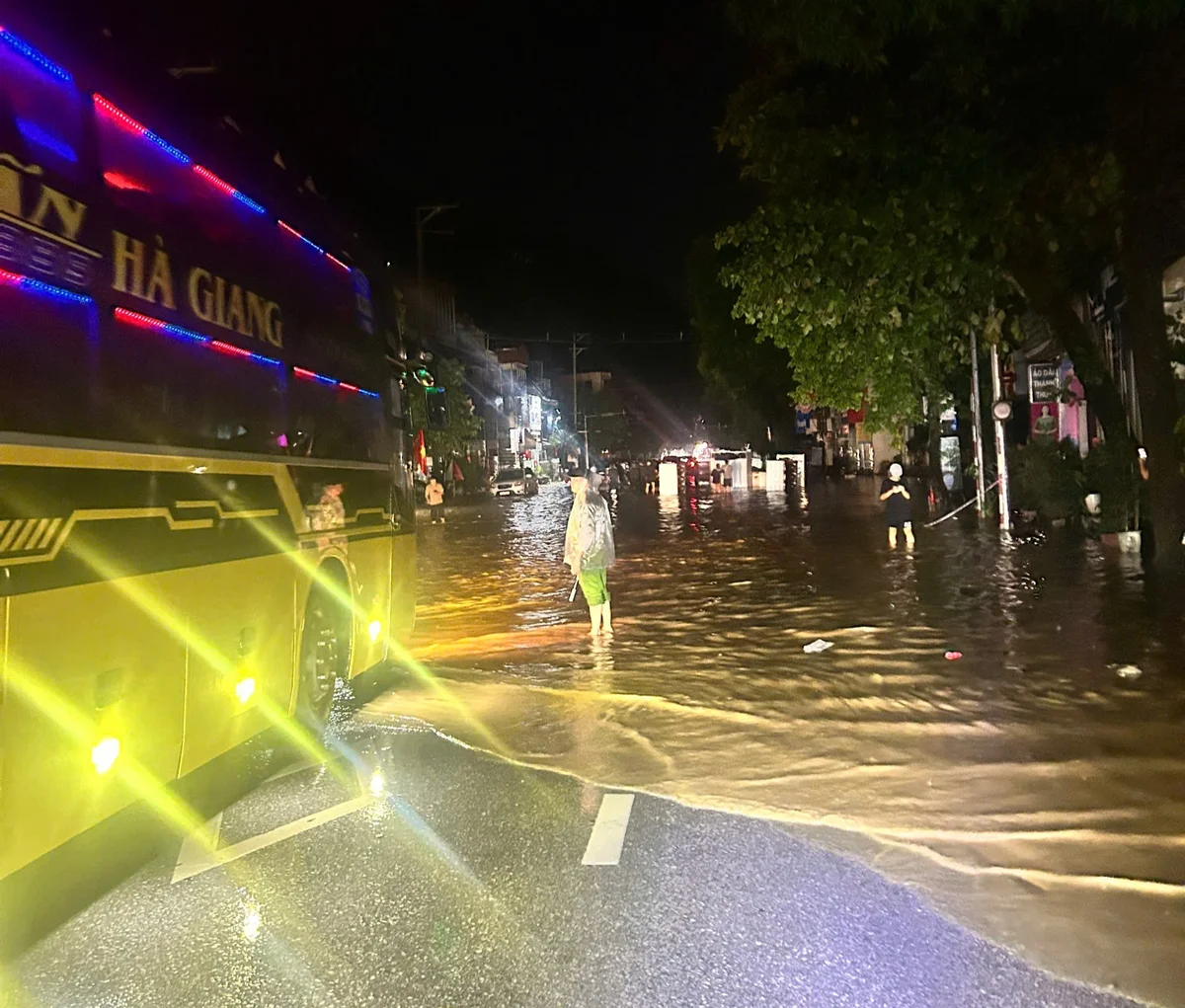 Nighttime flood scene with people wading in water. A bus with neon lights is nearby. Street lights reflect on water. Mood is tense.