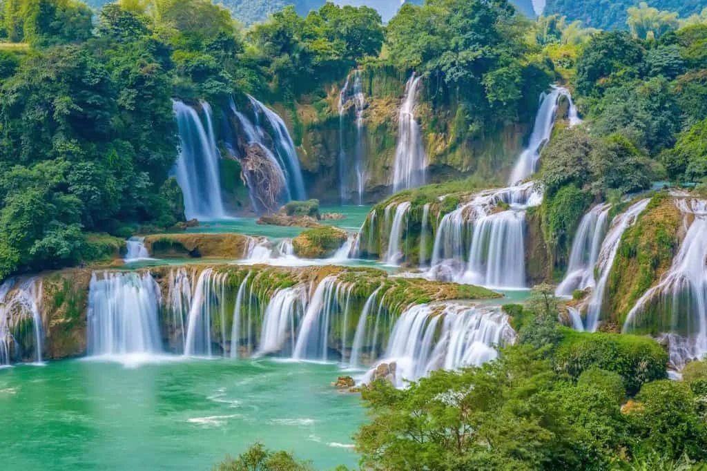 Lush green landscape with cascading waterfalls flowing into turquoise pools. Dense trees surround the falls, creating a serene, natural setting.