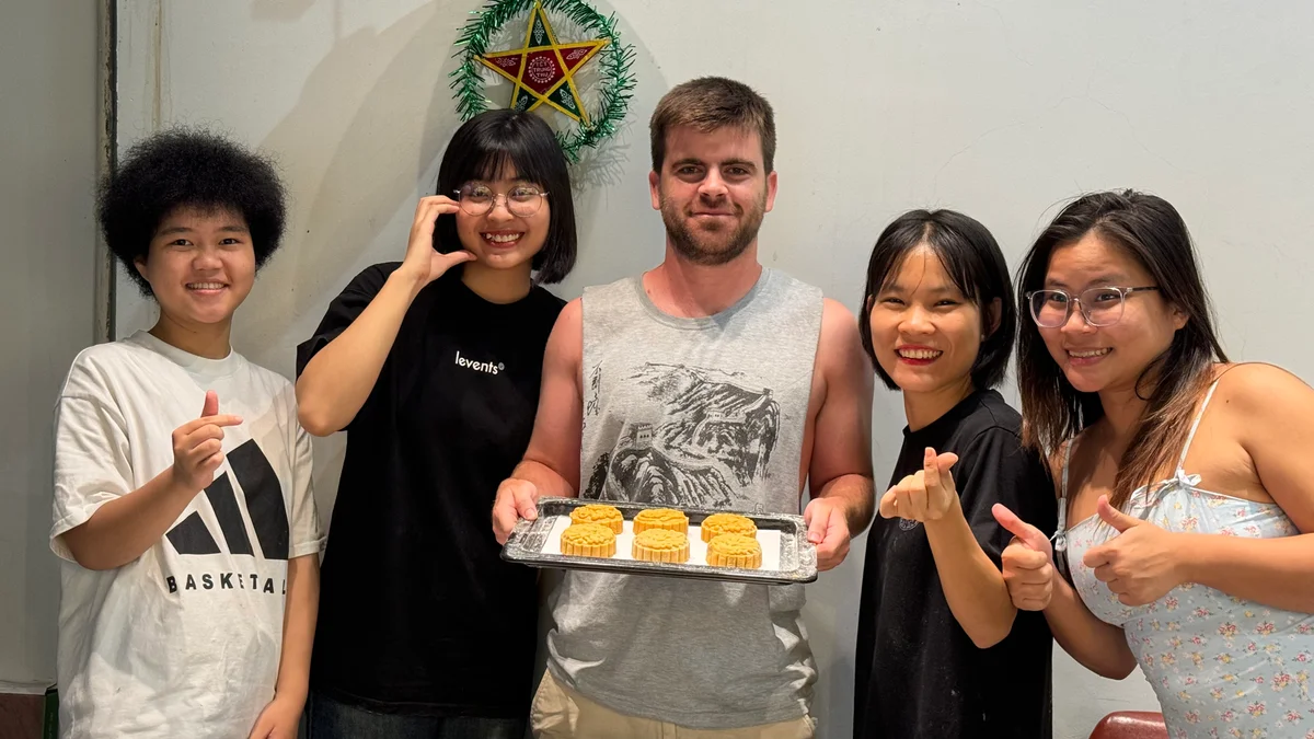 Five people smiling, holding a tray of mooncakes indoors. A star decoration hangs on the wall. Casual summer attire, cheerful mood.