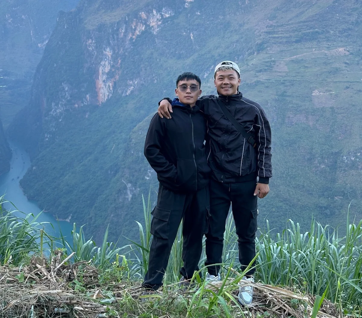 Two people in black jackets smile, standing on a cliff with lush green grass. A dramatic valley with a river lies in the background.