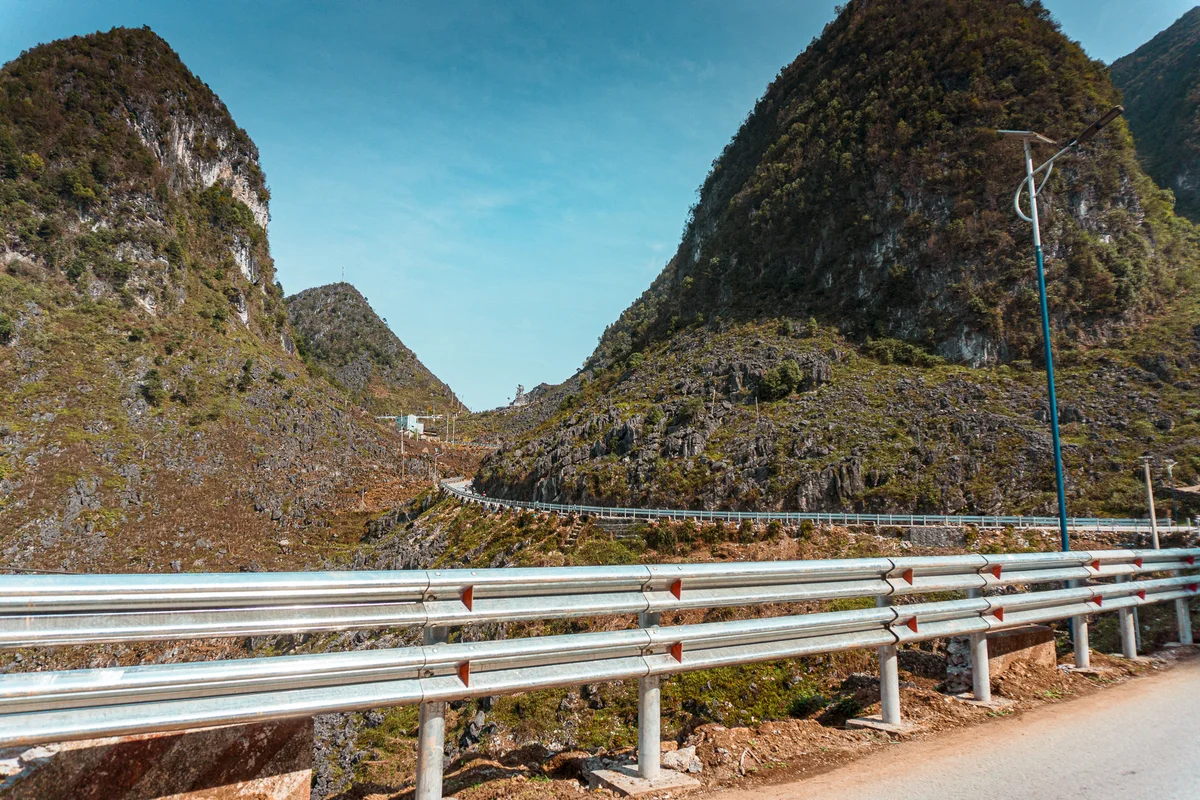 Winding road between lush green mountains under blue sky, bordered by metal guardrails. The mood is serene and scenic.