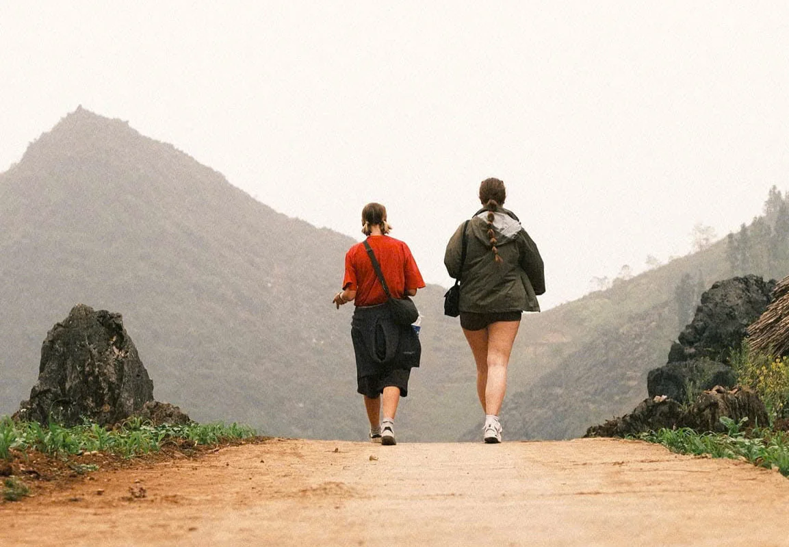 Two people walking on a dirt path in a mountainous area. One wears a red shirt, the other a green jacket. Overcast sky, rocky terrain.