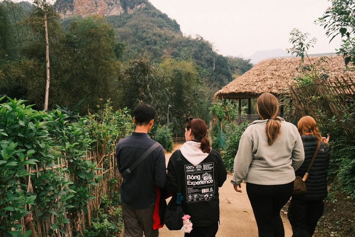 Four people walk on a path through lush greenery, near a straw-roofed hut. One wears a "Bòng Experience" jacket. Mountains in the background.