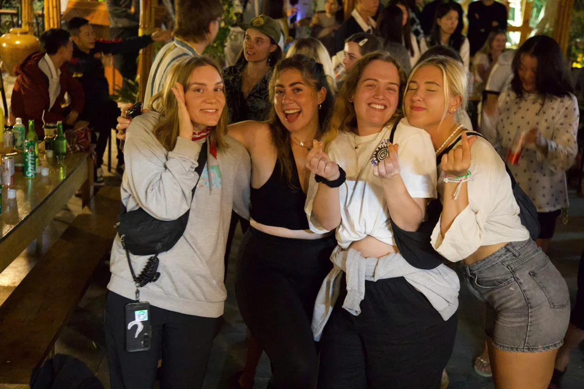 Four smiling women pose closely in a lively, crowded indoor setting. Drinks and casual attire suggest a fun, relaxed atmosphere.