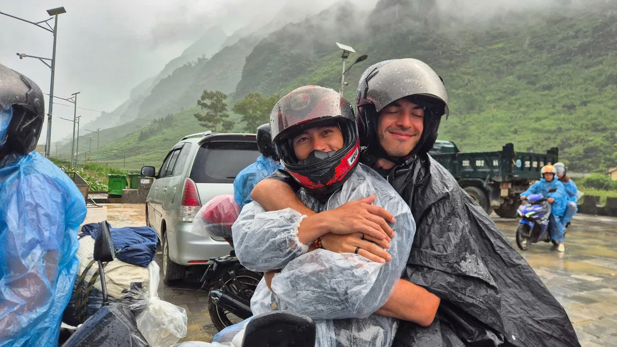 Two people in helmets and rain ponchos hug, smiling on a rainy day. Mountains and vehicles are in the misty background.