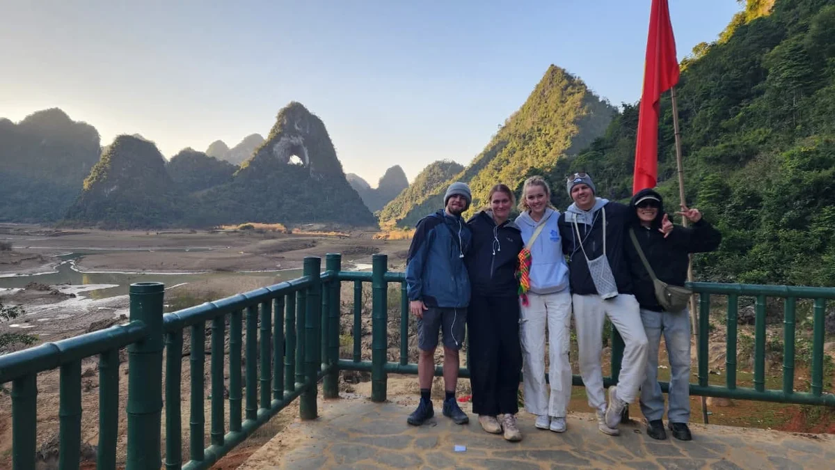 Group of five people smiling on a balcony with a red flag, scenic mountain landscape and riverbed in the background, clear sky overhead.