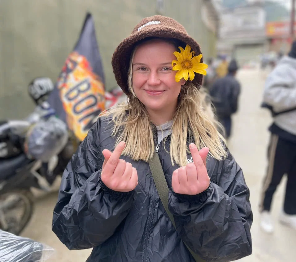A woman smiles, making a finger heart gesture, wearing a brown hat with a yellow flower in her hair. Blurry street scene in background.