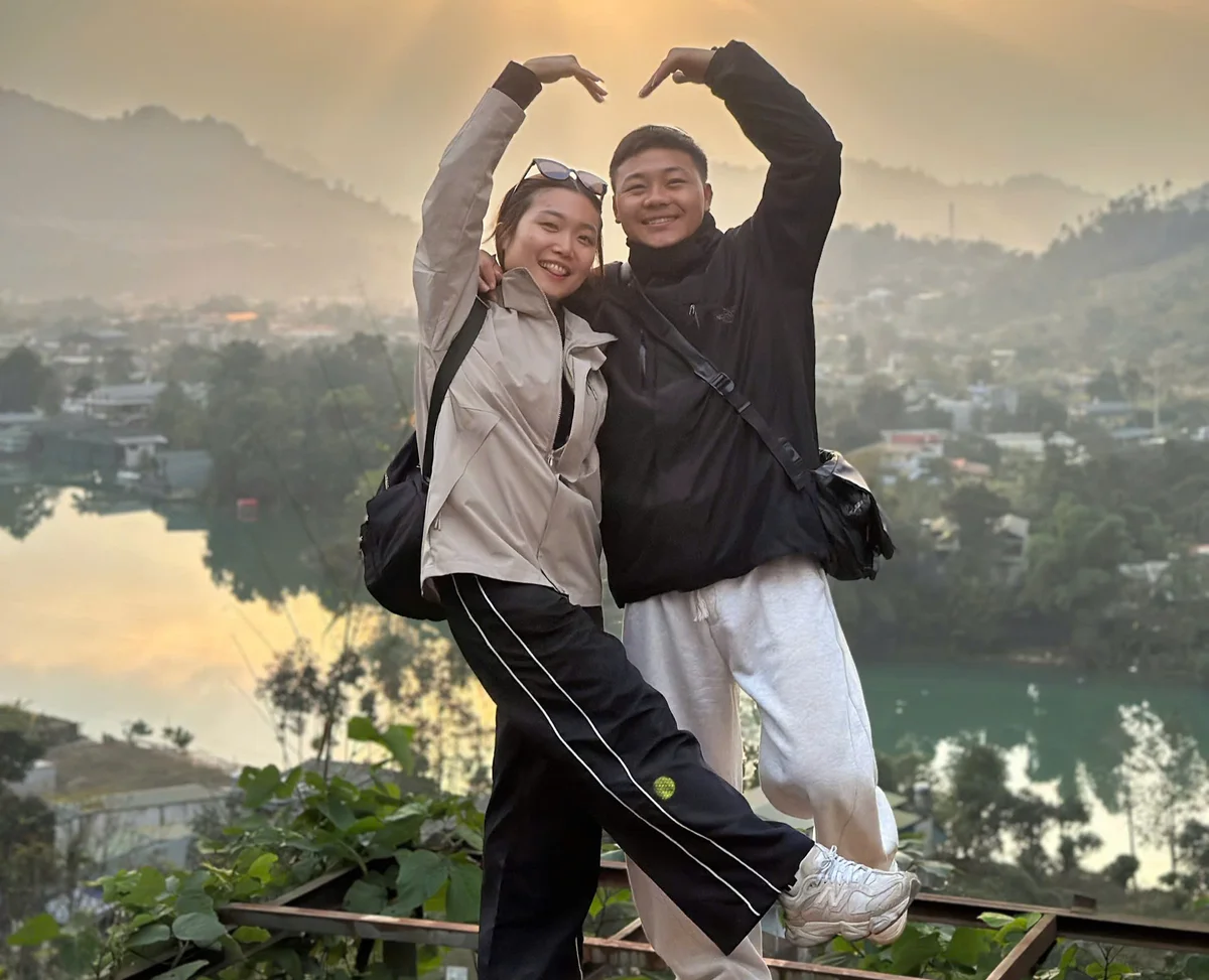 Two people pose with arms in heart shape, smiling. They're in casual clothes with a scenic view of hills and a lake at sunset in the background.