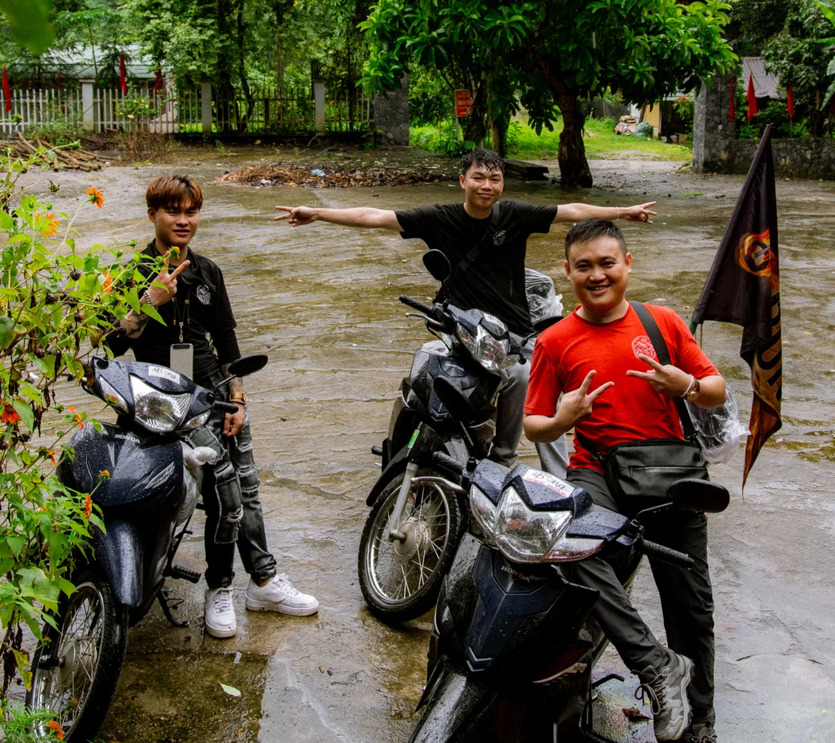 Three smiling men pose with motorcycles in a rainy, lush setting. One wears a red shirt and backpack, while two point with one hand each.