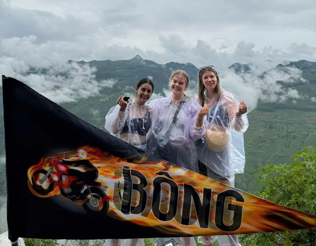Three women in clear ponchos smiling, holding a flag with a flaming motorcycle and "BONG" text, with cloudy mountains in the background.