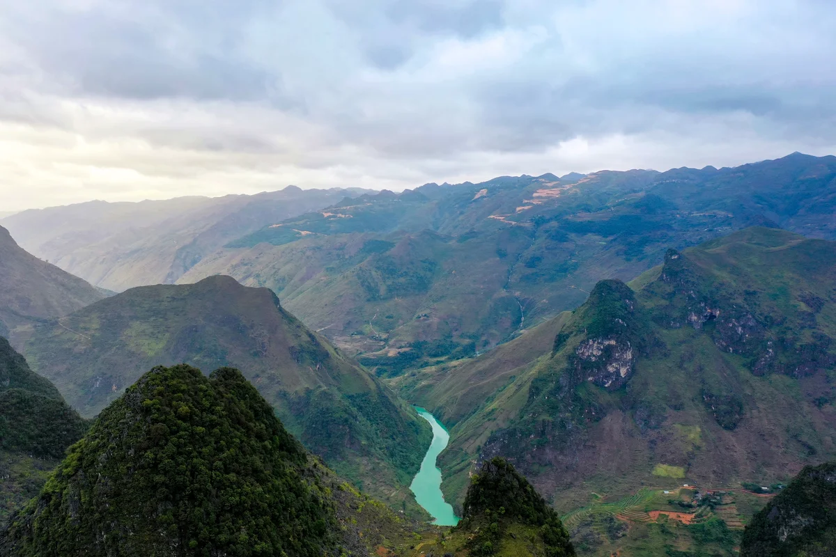 A winding turquoise river flows through lush green mountains under a cloudy sky, creating a serene and expansive landscape view.