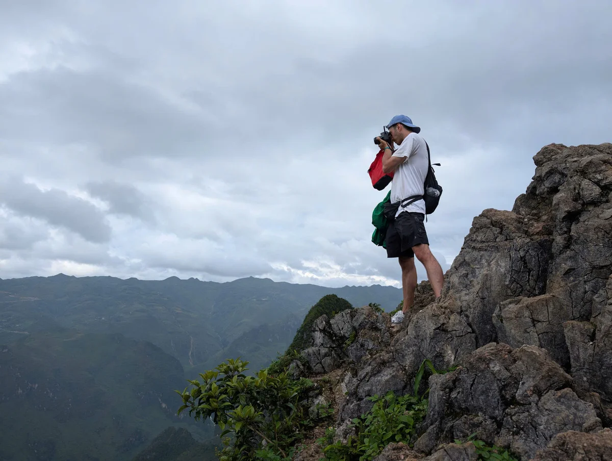 A person in a white shirt and blue cap photographs a mountainous landscape from rocky terrain under a cloudy sky, exuding adventure.