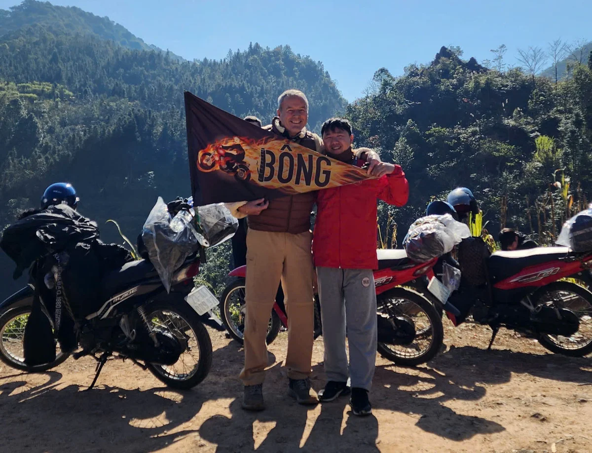 Two people pose with a "Bong" flag in a mountainous area. Motorbikes are beside them, and they're smiling against a clear blue sky.