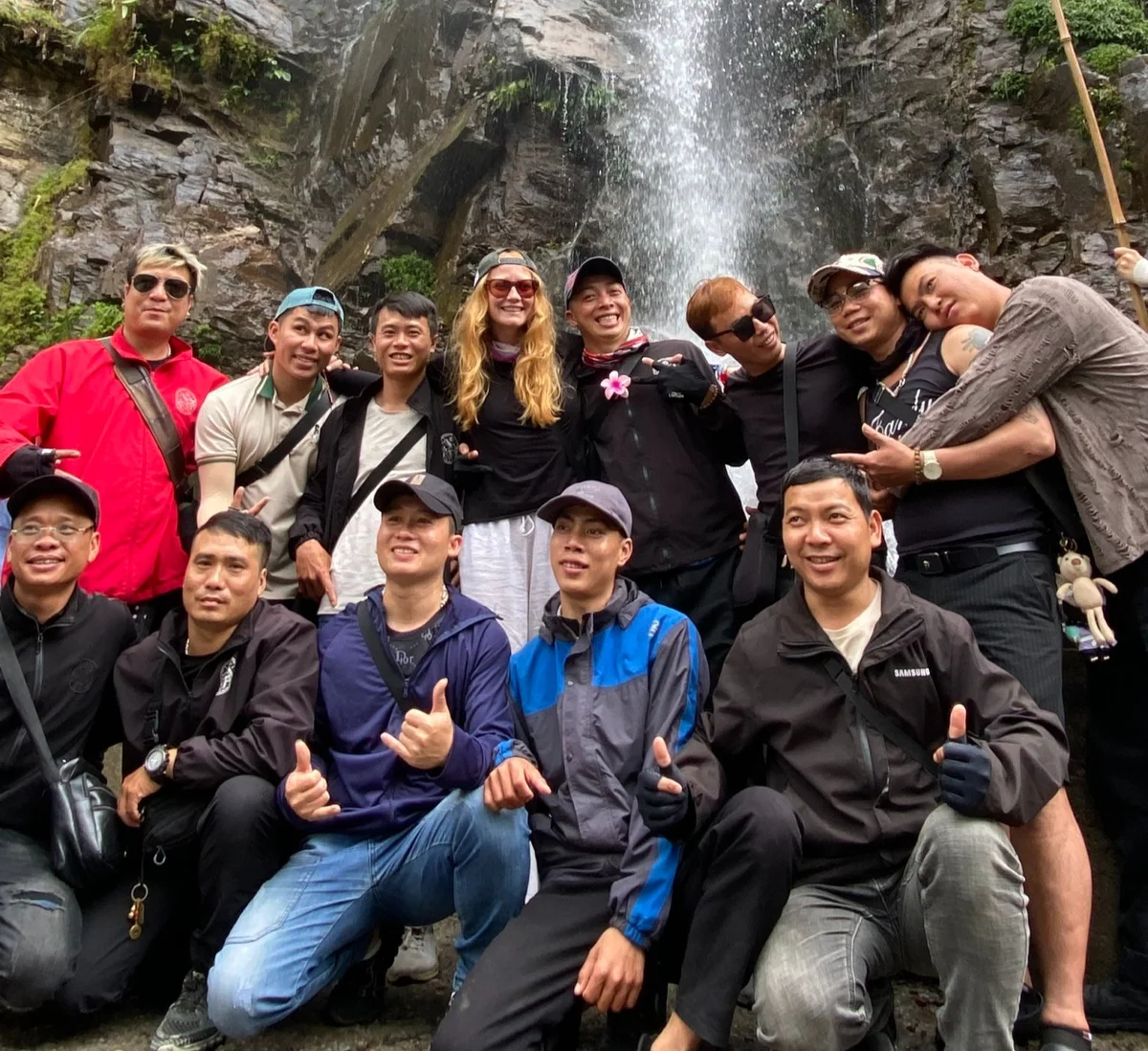 A group of people pose happily in front of a waterfall, wearing casual outdoor clothing. One person wears a bright red jacket. Rocks and greenery in background.