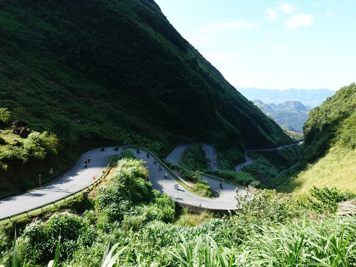 A winding mountain road with several motorbikes, surrounded by lush green hills under a clear blue sky, creating a serene, vibrant scene.