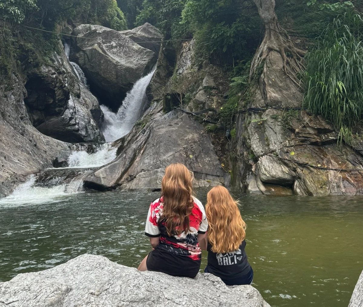 Two people with long hair sit on a rock, facing a cascading waterfall in a lush, green forest setting. Tranquil and scenic mood.