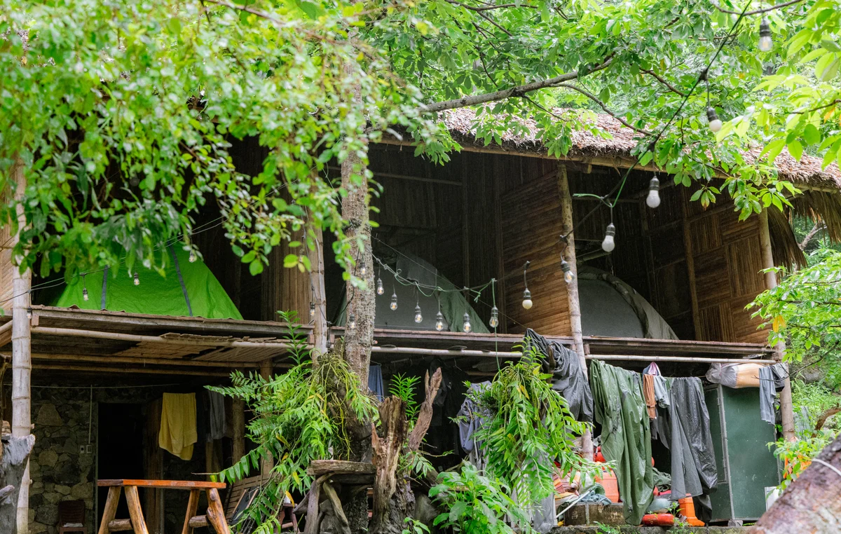 Rustic treehouse with green tent on wooden balcony, surrounded by lush foliage and string lights. Clothes hang to dry outdoors.