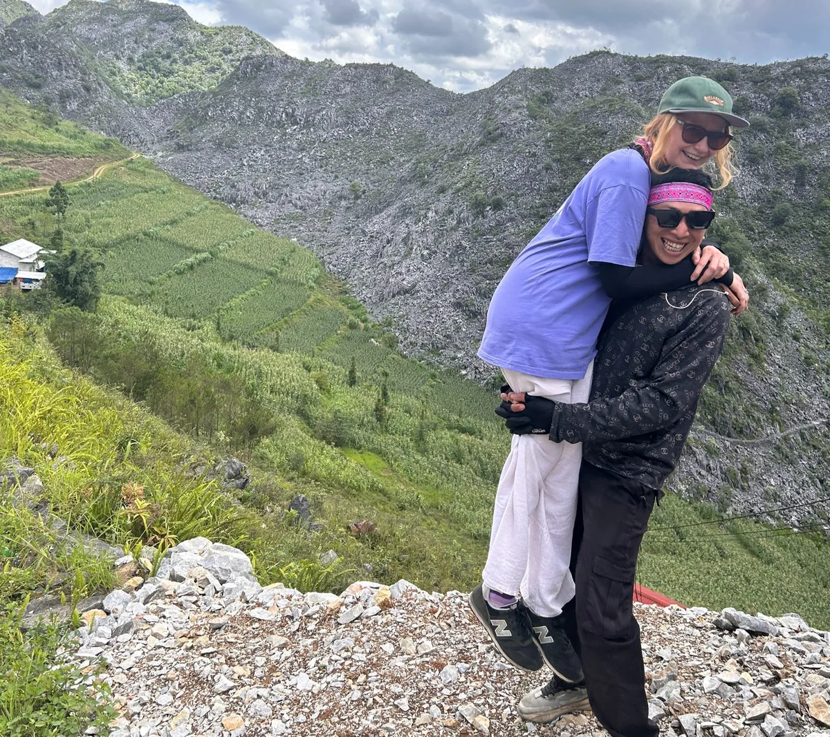 Two people happily pose on a rocky hill with lush green fields and mountains in the background. The mood is joyful and adventurous.