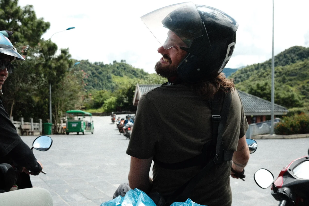 Man wearing helmet smiles on a motorcycle, mountainous backdrop with trees and distant scooters. Blue plastic bag visible. Vibrant mood.