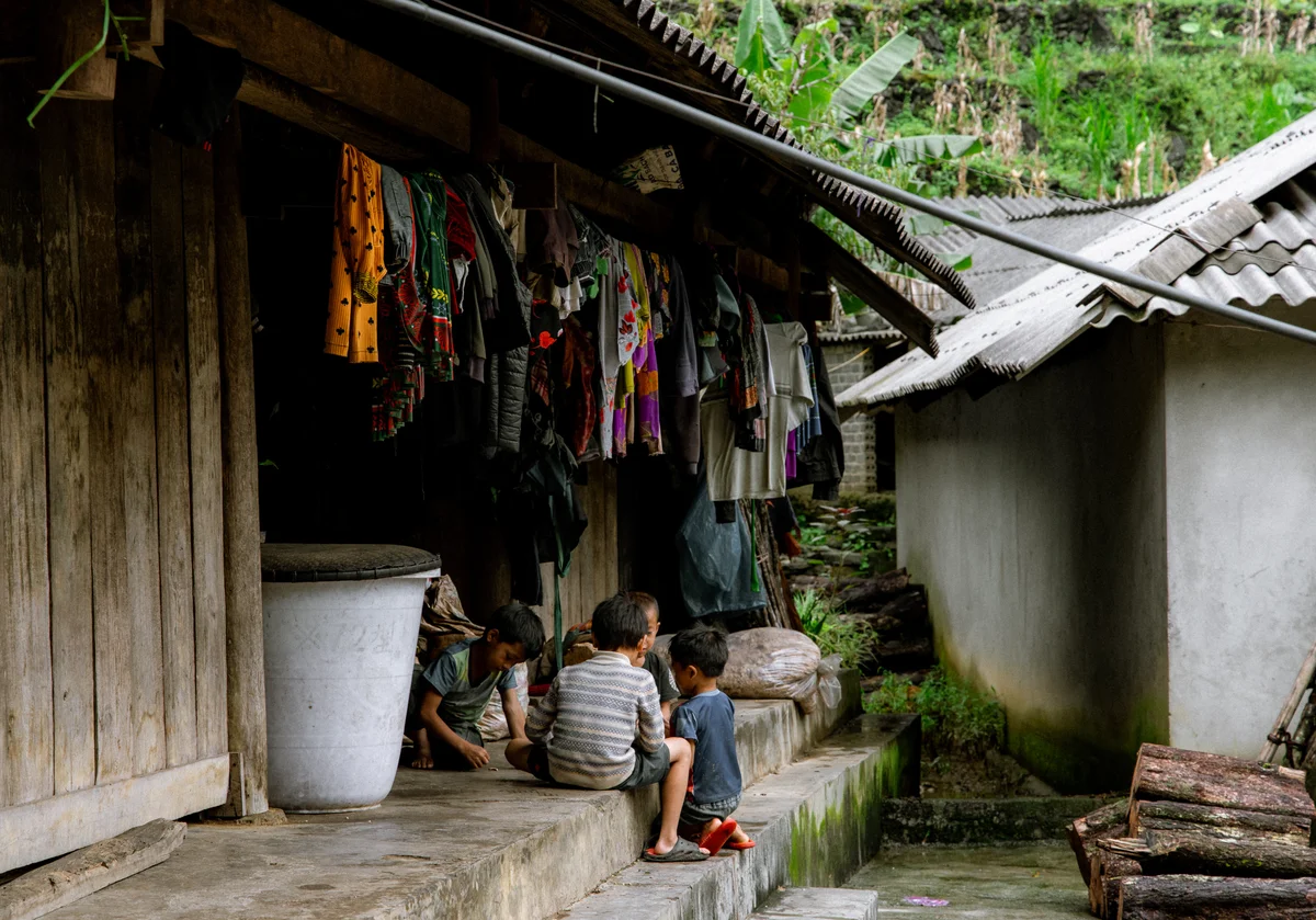 Four children sit and play on a concrete ledge outside a wooden building with colorful clothes hanging, surrounded by lush greenery.