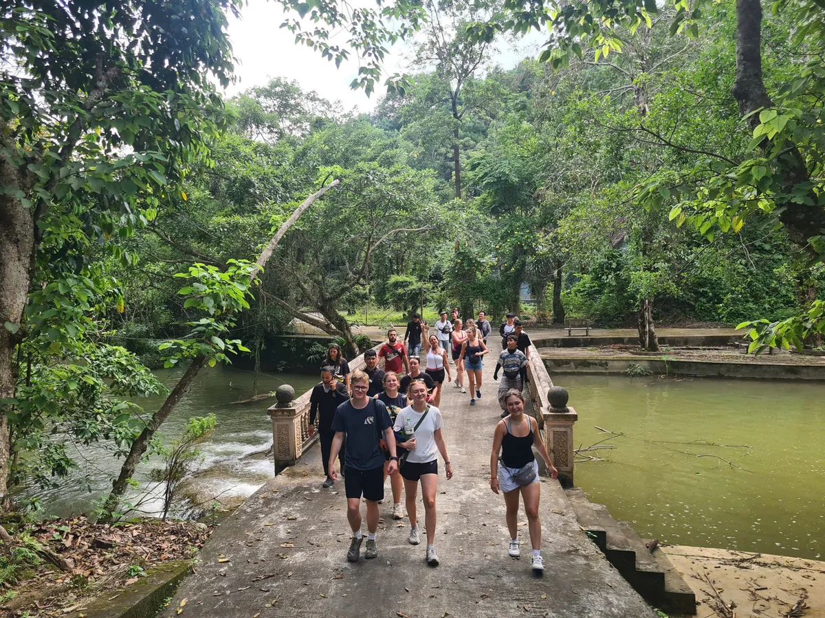 Group of people walking on a stone bridge in a lush green forest. River flows beneath. Overcast sky, casual attire, cheerful mood.