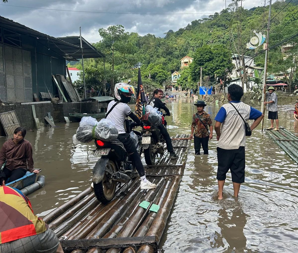 People using makeshift bamboo rafts to transport motorcycles across a flooded street. Overcast sky and green trees in the background.