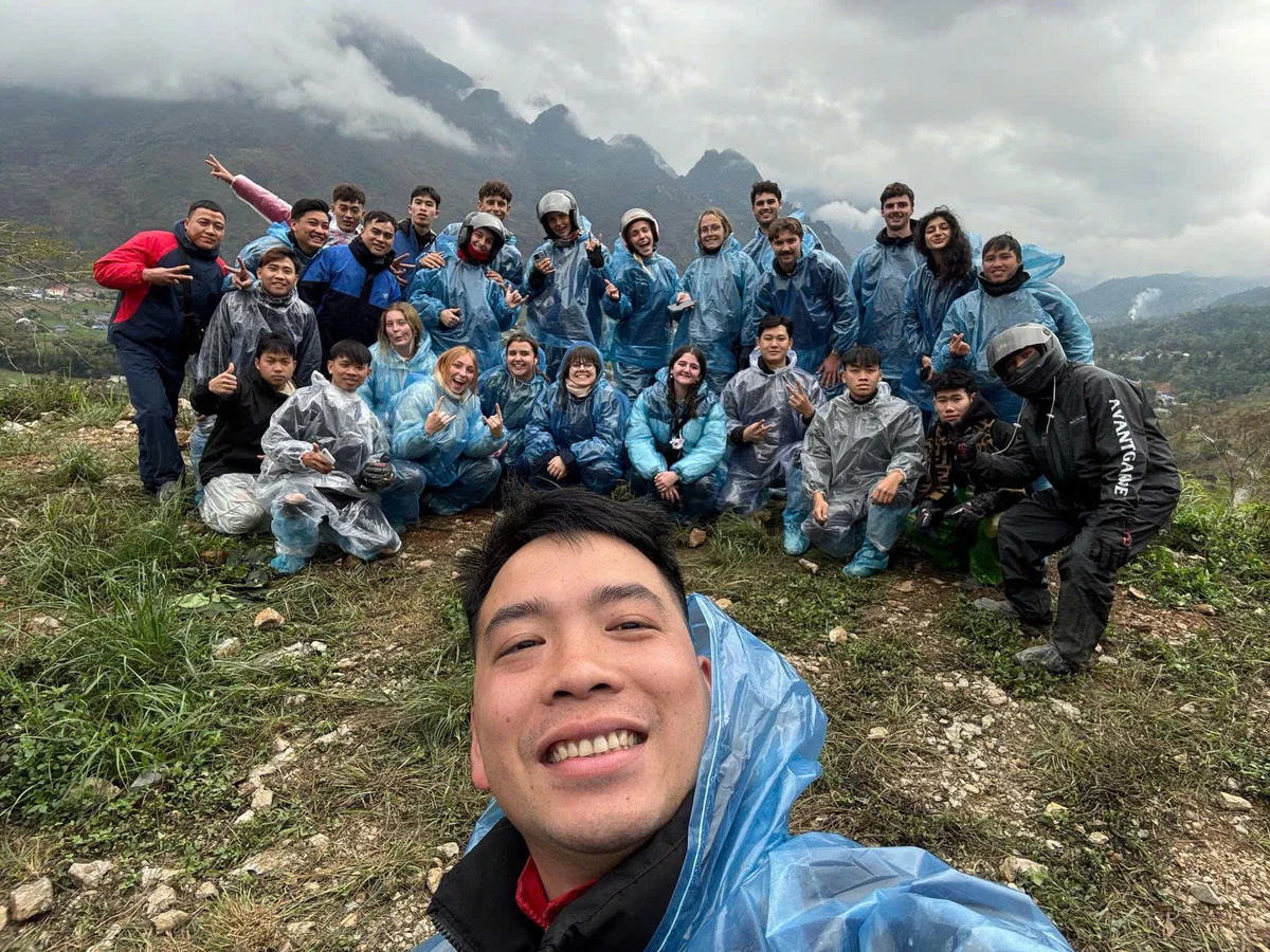 Group of people in blue rain ponchos pose for a selfie on a misty mountain landscape, smiling and showing peace signs. Cloudy mood.