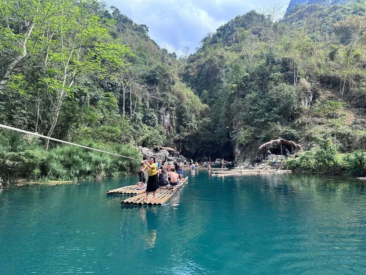 People rafting on a bamboo raft in a turquoise river, surrounded by lush greenery and rocky cliffs, convey a peaceful, adventurous mood.