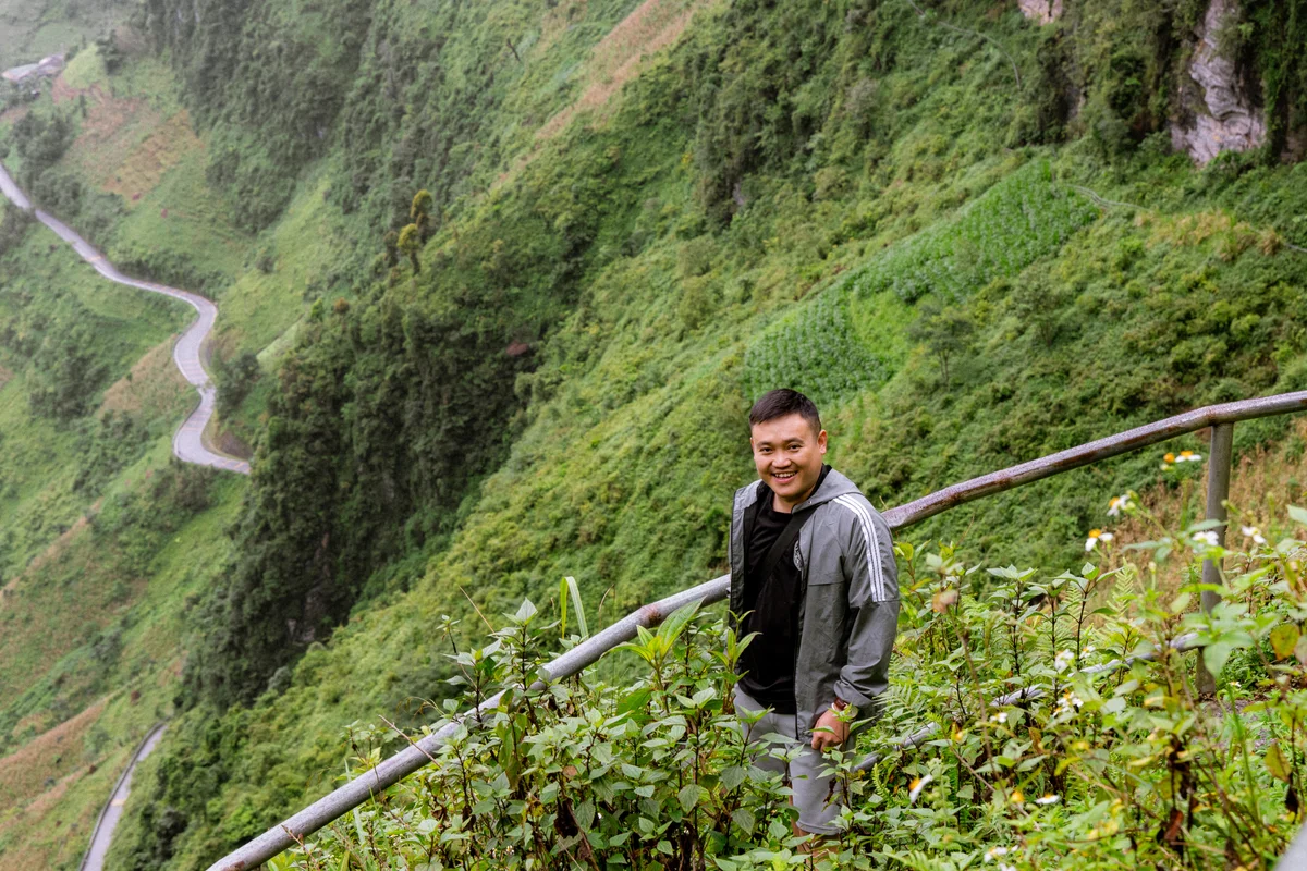 Man smiling on a lush, green hillside with winding road behind, wearing a gray jacket. Bright, vibrant greenery adds a serene mood.