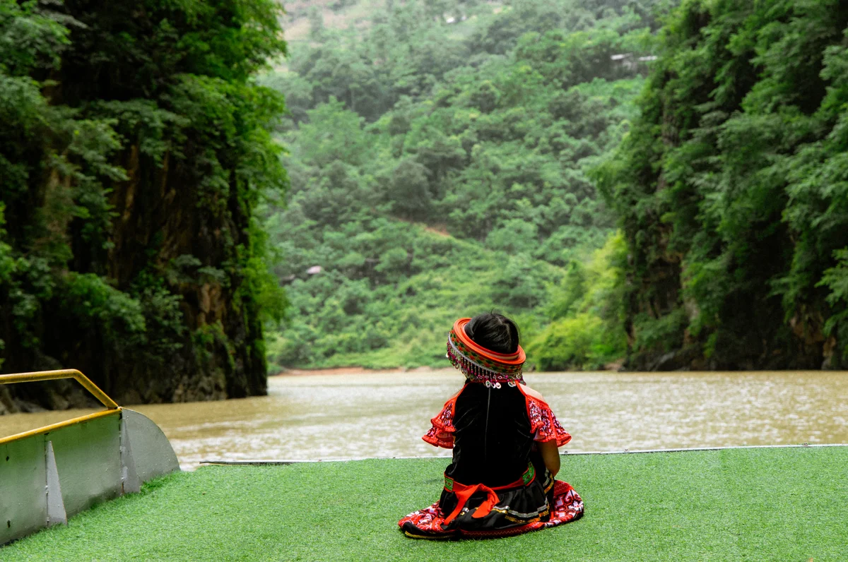Child in colorful attire sits facing a scenic river and lush green cliffs, evoking a sense of peace and contemplation, under a cloudy sky.