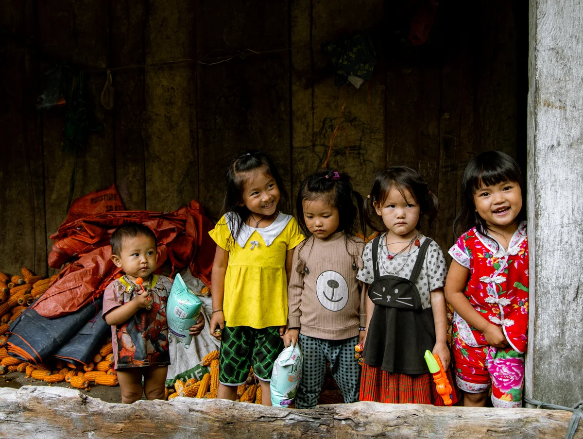 Five smiling children in colorful clothes stand in front of a wooden wall with sacks and corn. They hold bags and toys, exuding joy.