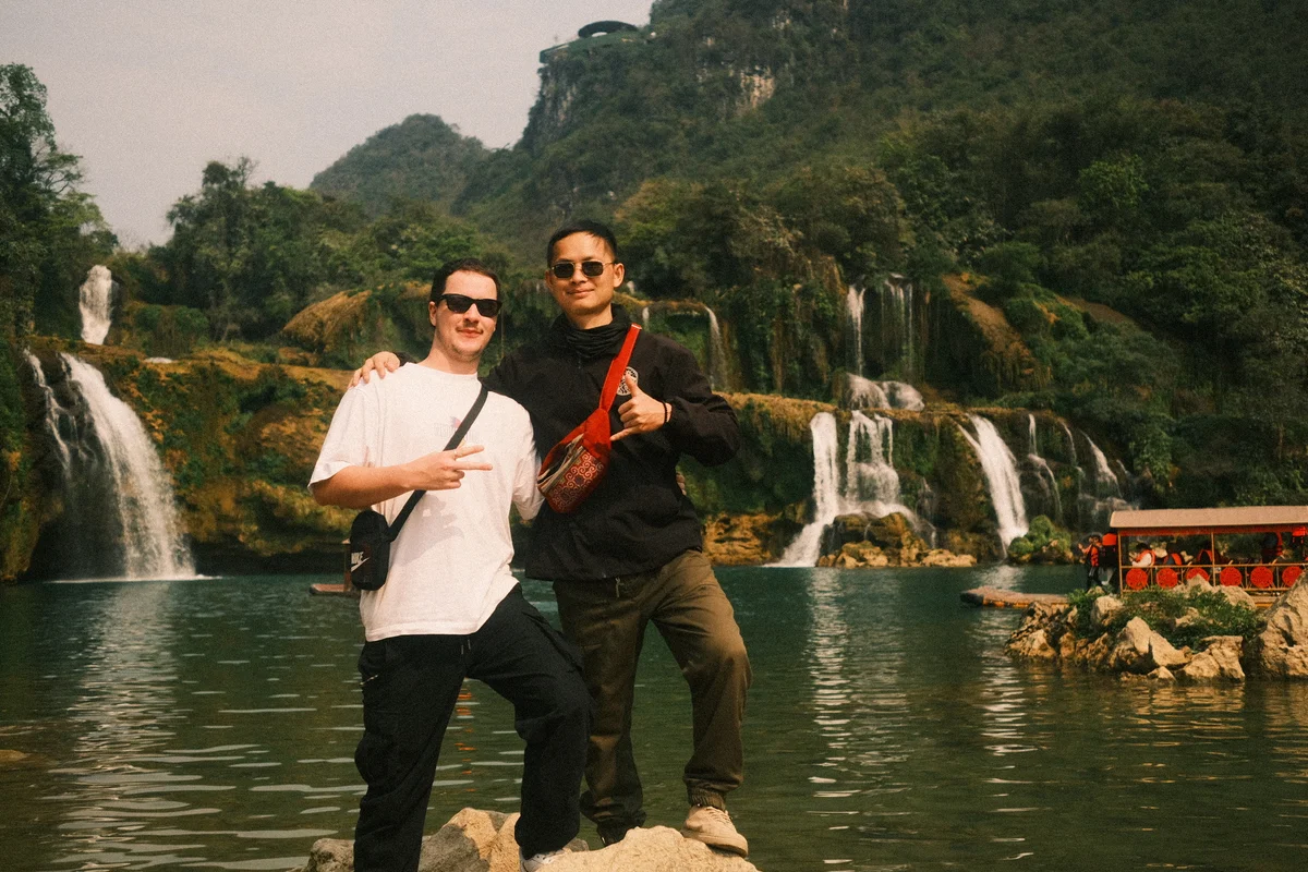 Two men pose happily at a scenic waterfall with lush greenery. One flashes a peace sign; the other gives a thumbs up. A boat is in the water.