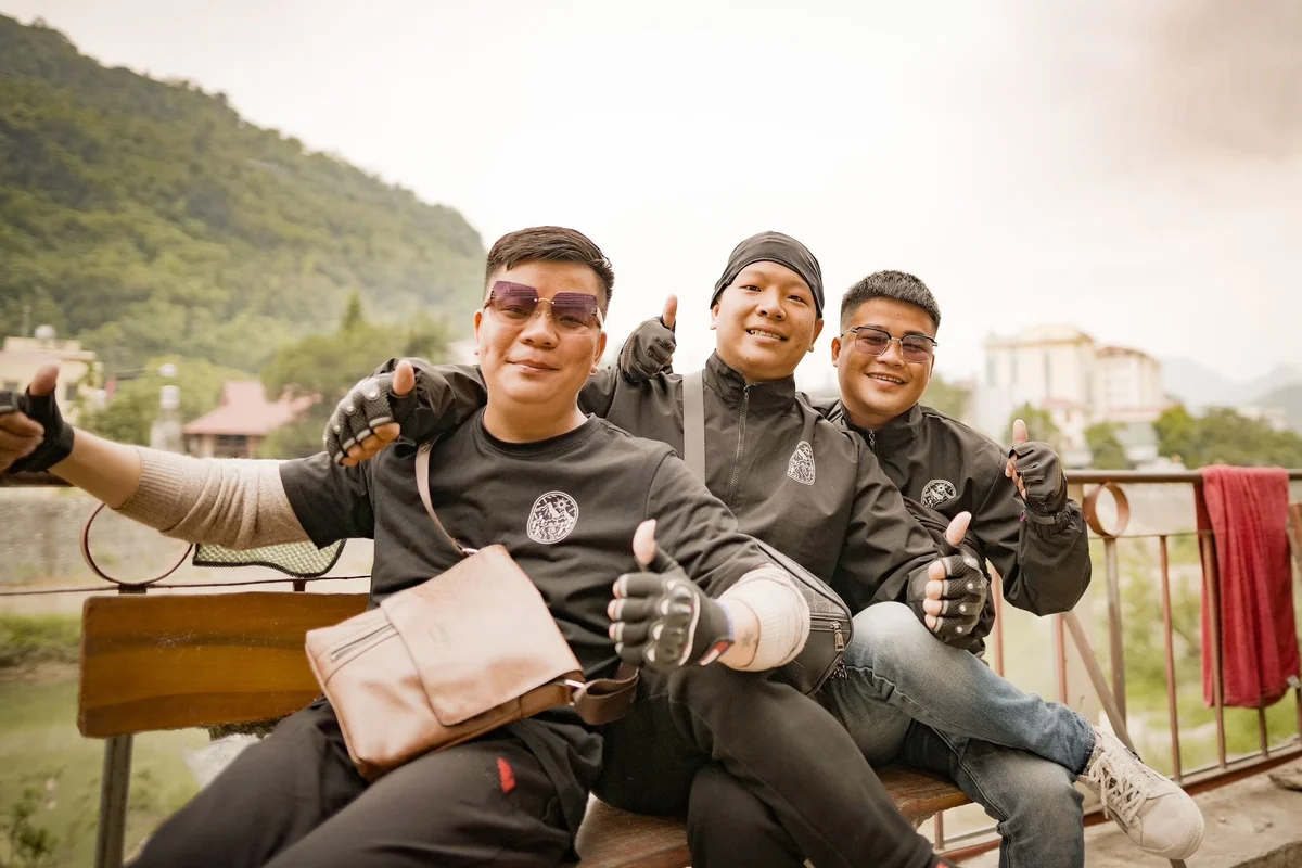 Three men in black jackets and gloves sit on a bench, smiling and giving thumbs up. Mountain and buildings in the background, relaxed vibe.